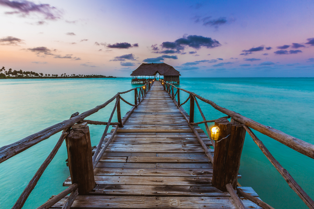 A long wooden foot bridge over an emerald beach that leads to an open native hut in the middle of the beach during sunset. 