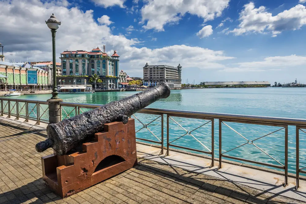 An old cannon displayed on the side of a boardwalk while visible on the other side are city structures. 