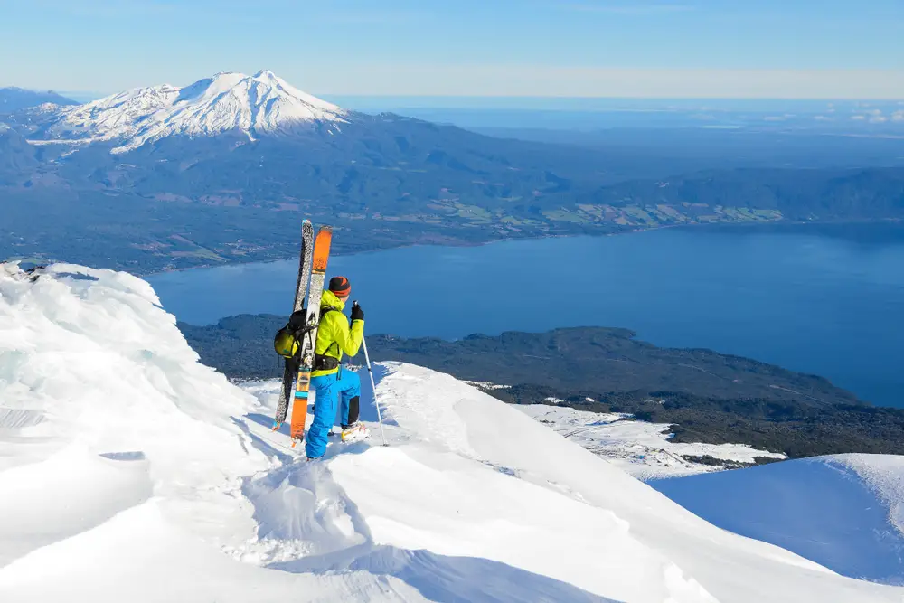 Ένας άντρας με κίτρινα και μπλε που ετοιμάζεται να κατέβει με σκι από την κορυφή ενός χιονισμένου βουνού και ένα άλλο ψηλό βουνό μπορεί να φανεί από μακριά με χιονοσκεπή. 