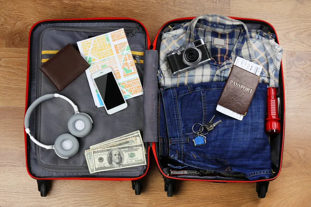 Packed, open suitcase prior to an international trip with money, passport, headphones, phone, map, and clothing visible in the shot taken from above