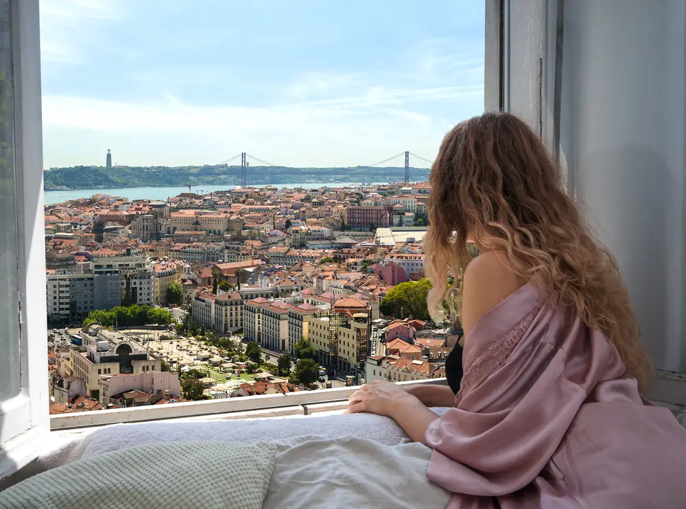 Μια γυναίκα σε ένα δωμάτιο ξενοδοχείου κοιτάζοντας το παράθυρο με θέα μια πόλη και μια μεγάλη γέφυρα.