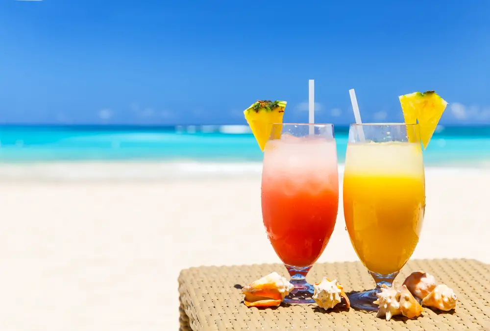 Δύο ποτήρια τροπικού ποτού με μια φέτα ανανά στην κορυφή του ποτηριού, τοποθετημένα σε ένα τραπέζι στην παραλία. 