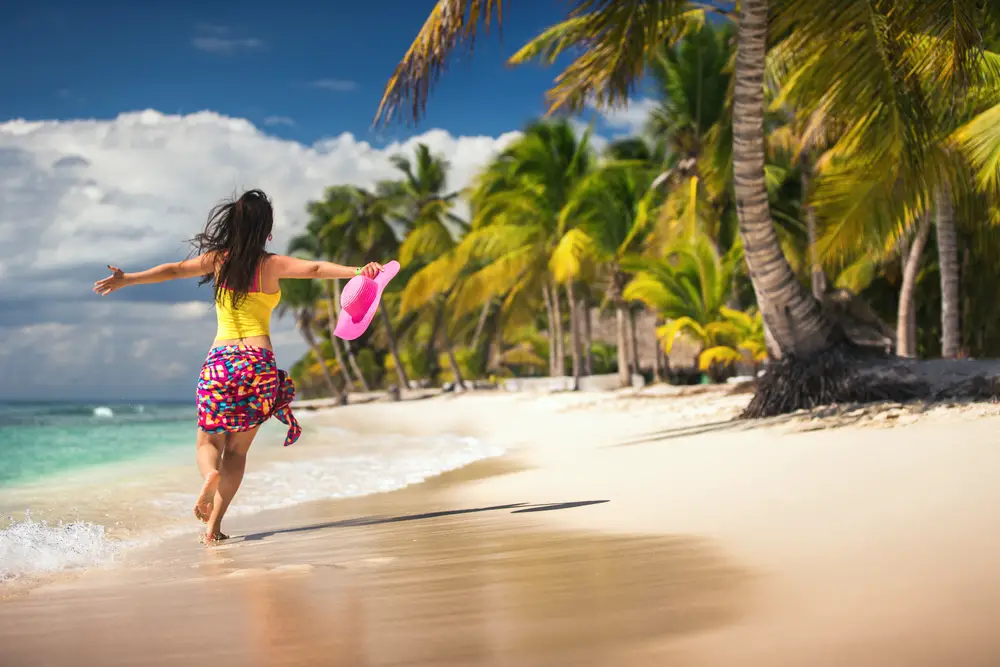 Μια ανέμελη γυναίκα που τρέχει στην παραλία με απλωμένα χέρια και κρατά το ροζ καπέλο της. 