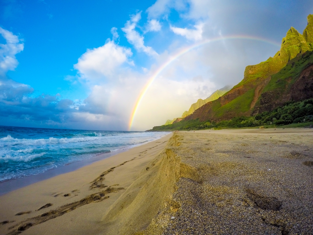 Θέα στην παραλία από την ακτή Napali στο Kauai, Χαβάη με ένα ουράνιο τόξο που αψιδώνει πάνω από την άμμο και τα ηφαιστειακά βουνά για ένα κομμάτι που απαντά σε ποια γλώσσα ομιλείται στη Χαβάη
