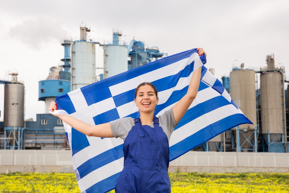 Μια γυναίκα εργάτρια εργοστασίου υψώνει την ελληνική σημαία ενώ το εργοστάσιο είναι πίσω της.