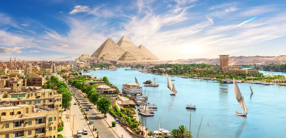The wide nile river with several boats in it, and the pyramid in background. 