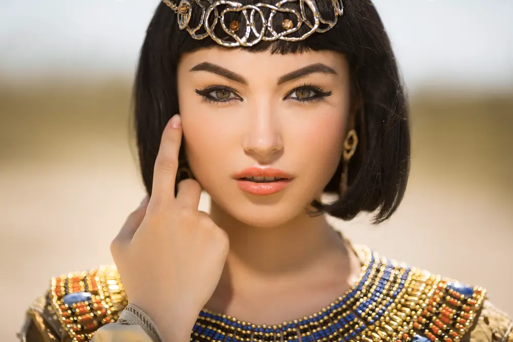 A woman wearing accessorised clothes pointing at her eyes with heavy makeup.