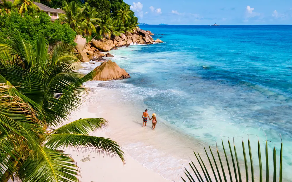 Ένα ζευγάρι που κρατιέται χέρι-χέρι απολαμβάνοντας την παραλία με ψιλή άμμο και μεγάλους βράχους. 