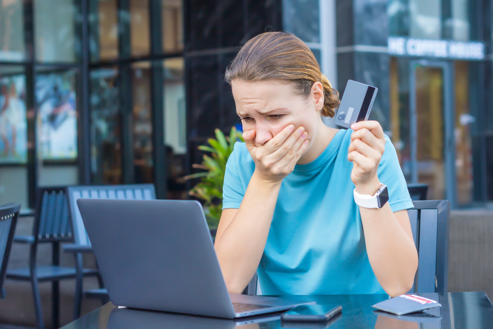A woman shocked and saddened while looking at her laptop monitor and holding her credit card. 