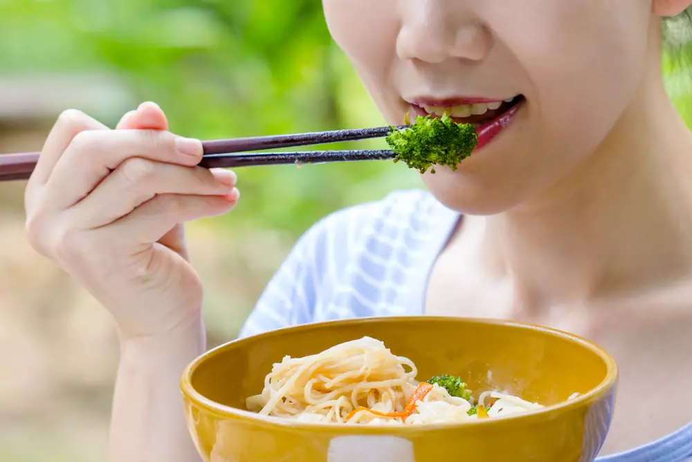 A woman eating a food bowl with pasta and broccoli using chopsticks.