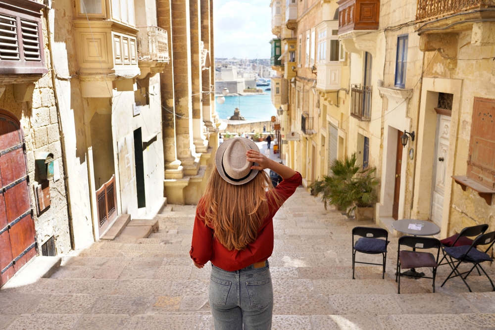 A women wearing red blouse holding her hat over her head while looking ahead a small alley. 
