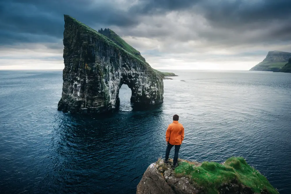 A man standing on the edge of a cliff looking at a cliff island in the middle of the sea. 