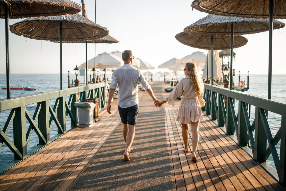 Ένα ζευγάρι που περπατά κρατώντας τα χέρια σε έναν πεζόδρομο με γηγενείς ομπρέλες και η θάλασσα είναι ορατή στο βάθος. 