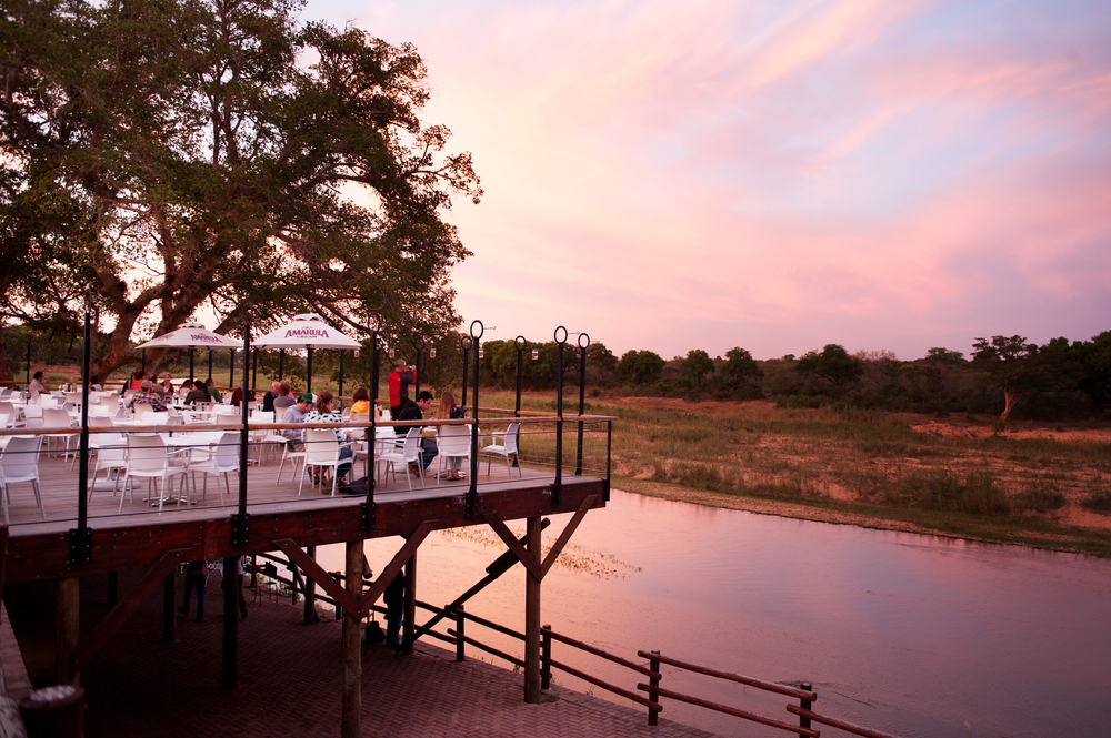 Ένα εστιατόριο χτισμένο σε μια υπερυψωμένη ξύλινη πλατφόρμα δίπλα σε ένα ποτάμι όπου μπορεί κανείς να δει κόσμο να τρώει κατά τη διάρκεια του ηλιοβασιλέματος.