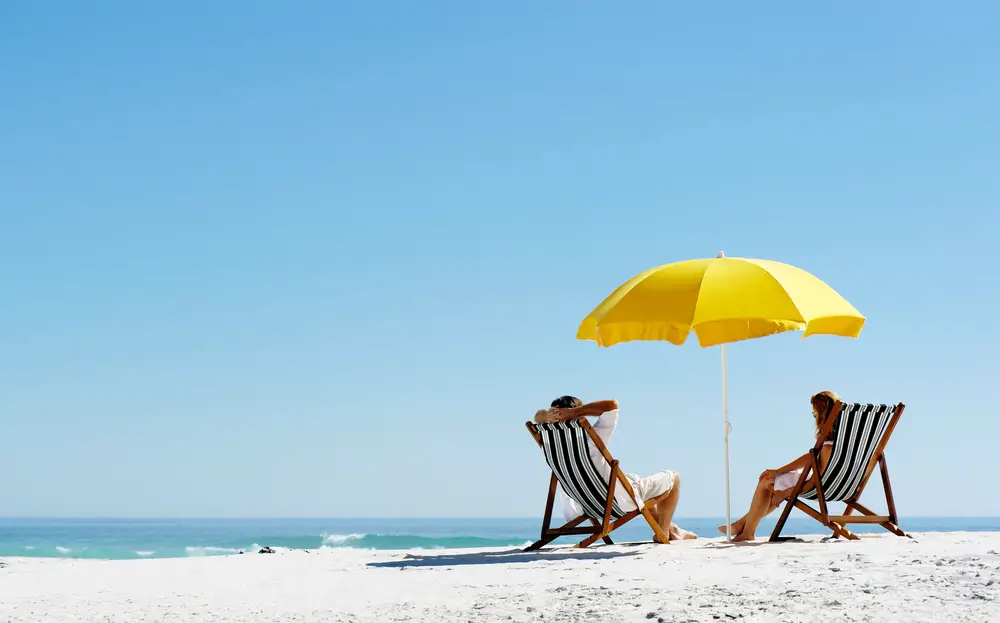 Ένα ζευγάρι που κάθεται σε ξαπλώστρες κάτω από μια κίτρινη ομπρέλα ενώ βρίσκεται σε μια παραλία με λευκή άμμο.