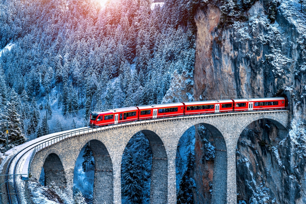Train passes along a bridge through the Alps in Switzerland with snowy winter scenery around for a guide on how to travel Europe by train