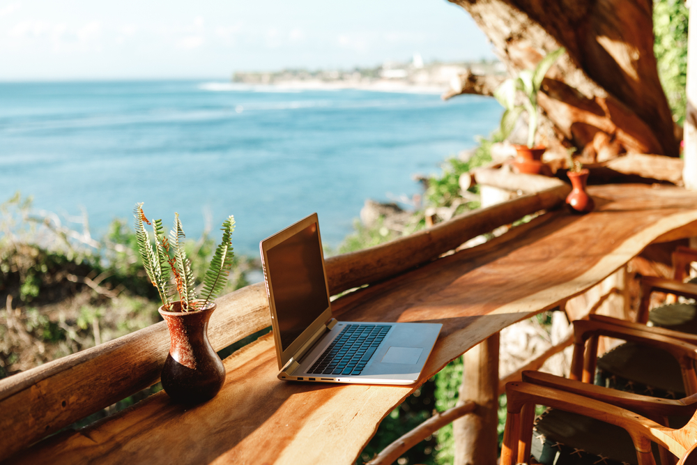 An open laptop sits on a wooden bar overlooking the ocean on a sunny day to indicate bleisure travel, or working and enjoying personal time on vacation
