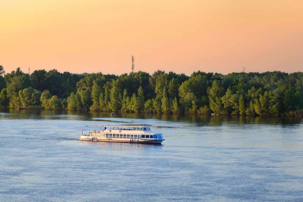 A cruise ship sailing on a wide river with trees on its banks during sunset. 