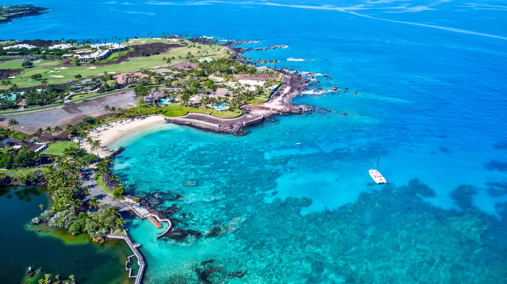 Aerial view of a section of the Big Island at Maunalani Beach Club with beautiful ocean surrounding the green edges and beaches for a guide showing how many people live in Hawai'i, the Big Island