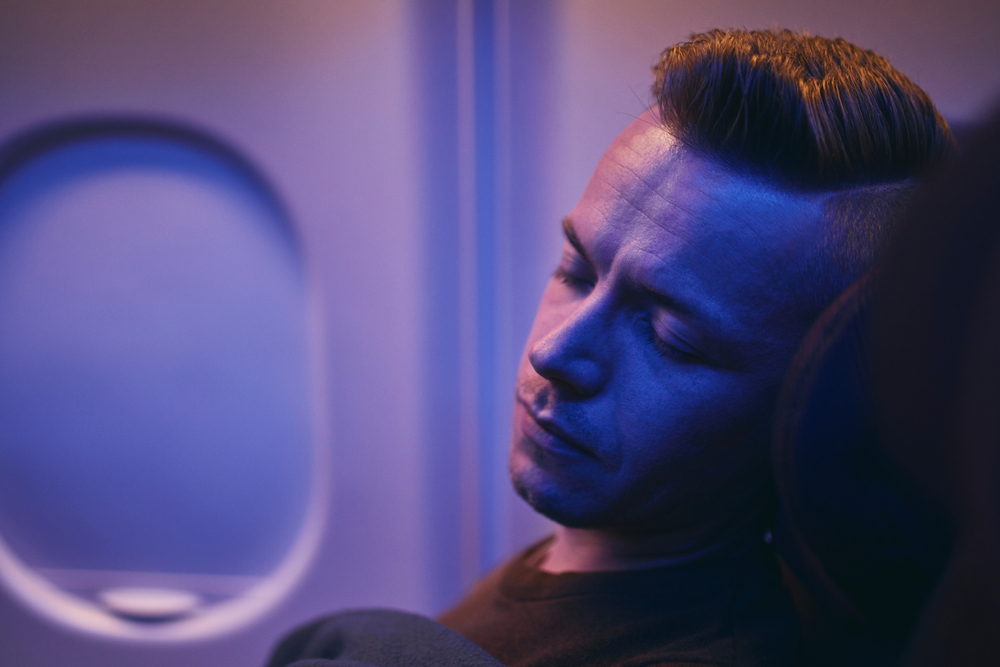 Άνδρας κοιμάται σε ένα αεροπλάνο κατά τη διάρκεια μιας πτήσης με κόκκινα μάτια με απαλό, μοβ χρώματος φως γύρω του 