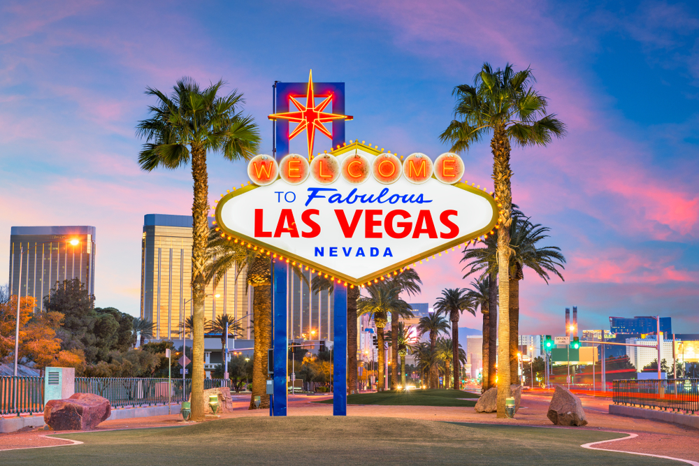 Άποψη της πινακίδας Welcome to Las Vegas στο Strip το σούρουπο που πλαισιώνεται από φοίνικες για μια ενότητα FAQ που περιγράφει λεπτομερώς πόσο διαρκεί μια πτήση προς Βέγκας