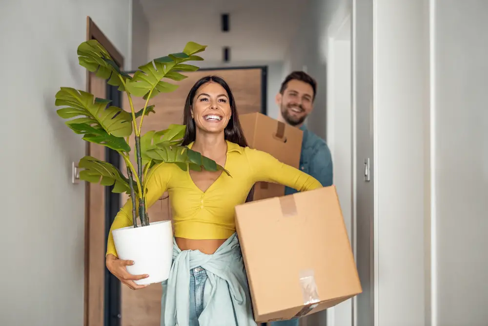Νεαρό ζευγάρι χαμογελά καθώς κουβαλάει κουτιά και φυτά εσωτερικού χώρου στο νέο τους σπίτι για να δείξουν την ιδέα του πώς να μετακομίσουν στη Χαβάη