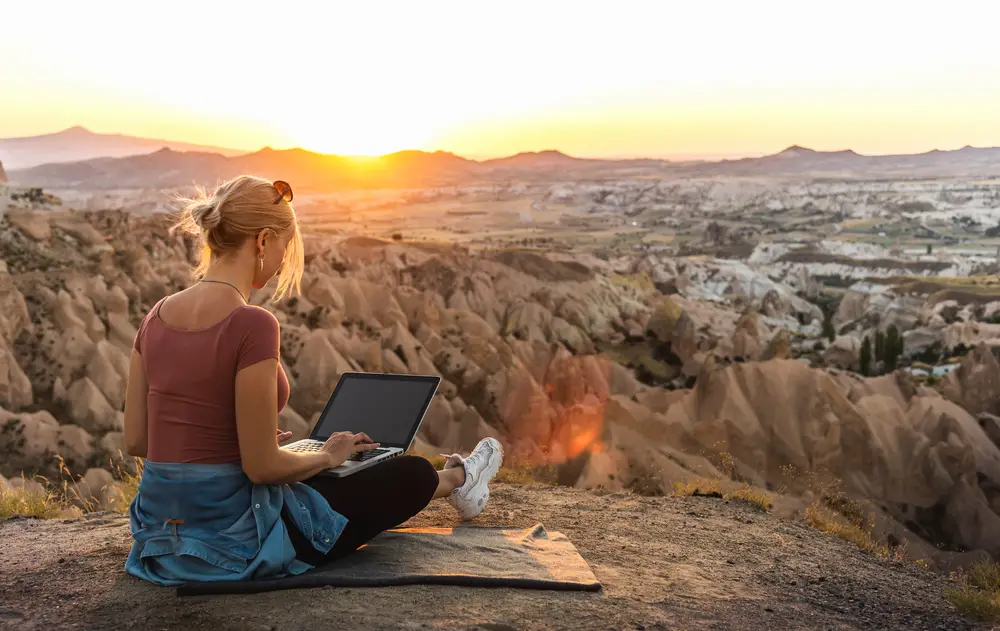 Concept of a digital nomad working on her laptop on a mountaintop at sunset for a guide detailing bleisure travel and how it works