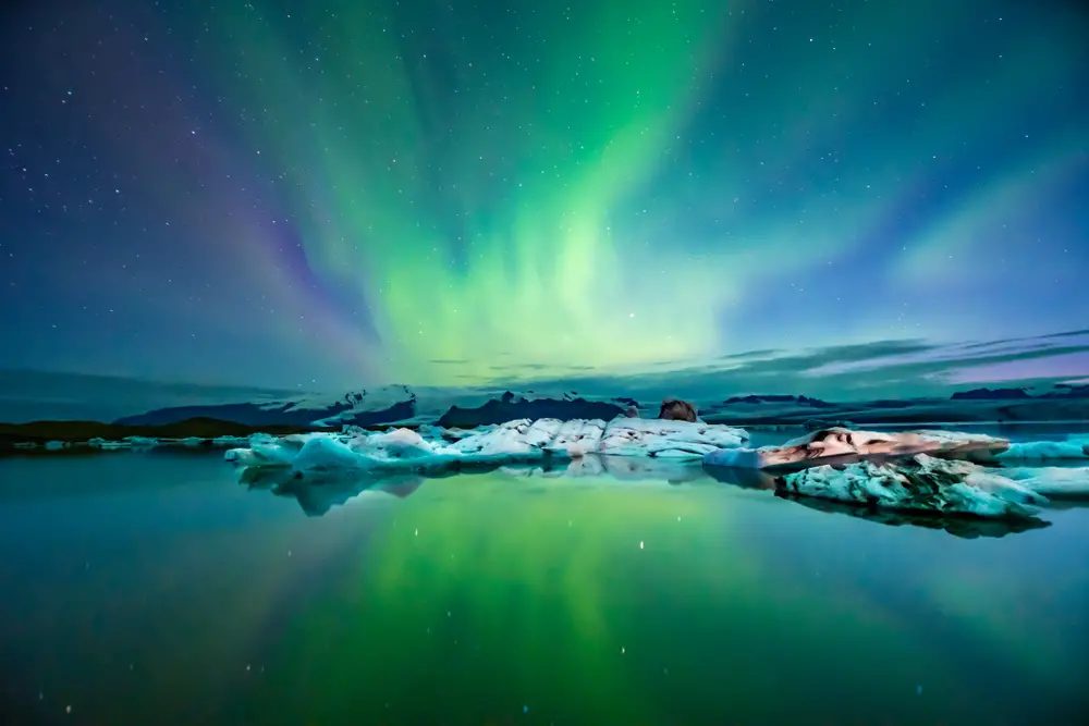 Η λιμνοθάλασσα Glacier στην Ισλανδία φαίνεται με πράσινο και μωβ Βόρειο Σέλας στον ουρανό πάνω τη νύχτα για έναν οδηγό που εξερευνά πόσο διαρκεί μια πτήση προς την Ισλανδία από τις ΗΠΑ