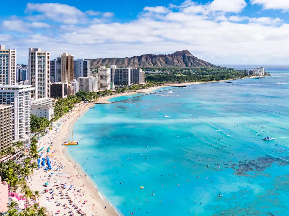Aerial view of Waikiki Beach on Oahu with Diamond Head crater in the distance as tourists line the beach for a guide demonstrating how many people live in Hawaii