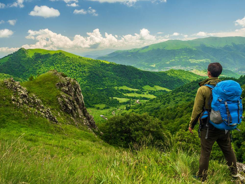 Ένας πεζοπόρος που κουβαλά ένα φορτωμένο μπλε σακίδιο πλάτης στέκεται στην πλαγιά ενός βουνού κοιτάζοντας μπροστά τα πράσινα βουνά.
