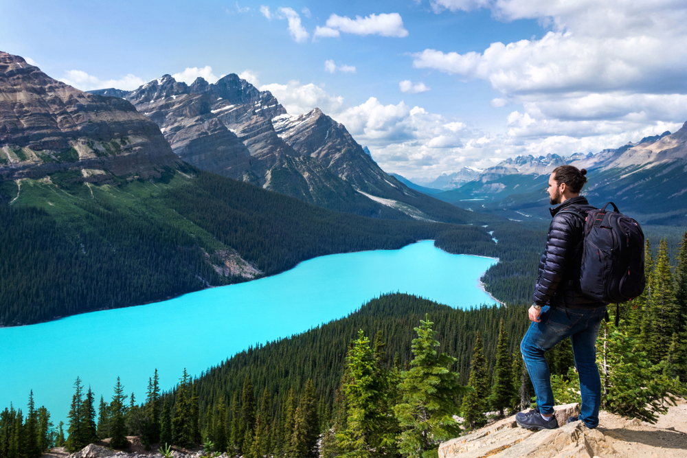 Ένας πεζοπόρος που στέκεται σε έναν βράχο με θέα τη θέα μιας λίμνης που αντανακλά τον ουρανό, τα βουνά και το δάσος από κάτω.