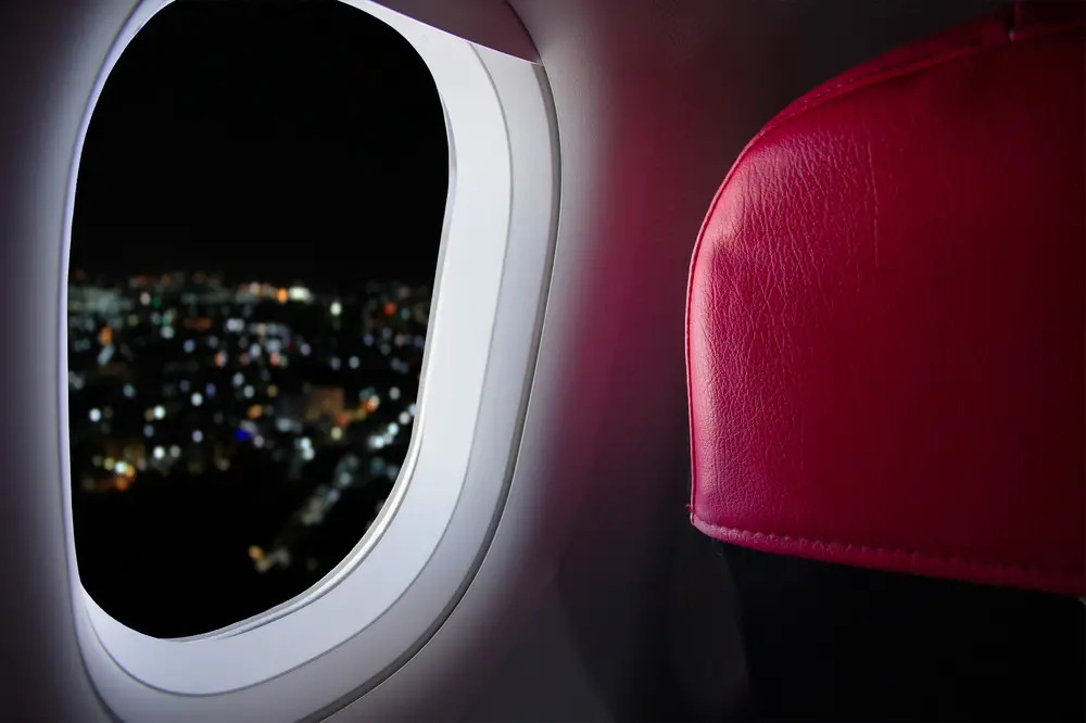 Παράθυρο επιβατών αεροπλάνου με κόκκινο προσκέφαλο καθίσματος που φαίνεται καθώς φώτα από την πόλη κάτω τη νύχτα κρυφοκοιτάγονται μέσα από το άνοιγμα για οδηγό για πτήσεις με κόκκινα μάτια