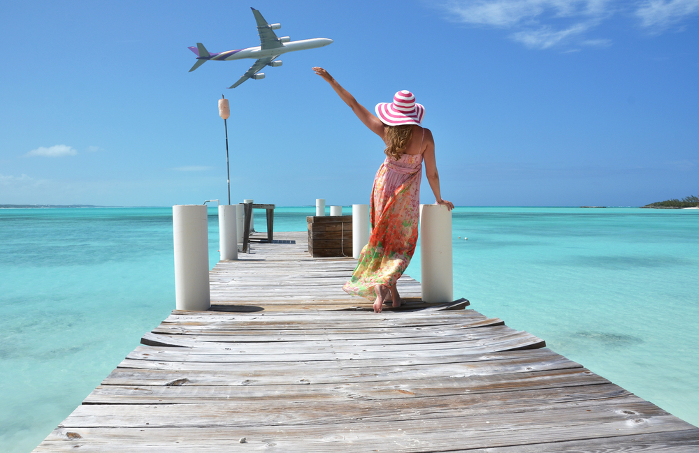 Μια γυναίκα που φορά ένα φόρεμα παραλίας ενώ στέκεται σε μια ξύλινη αποβάθρα, κουνώντας το χέρι της σε ένα διερχόμενο αεροπλάνο από πάνω. 