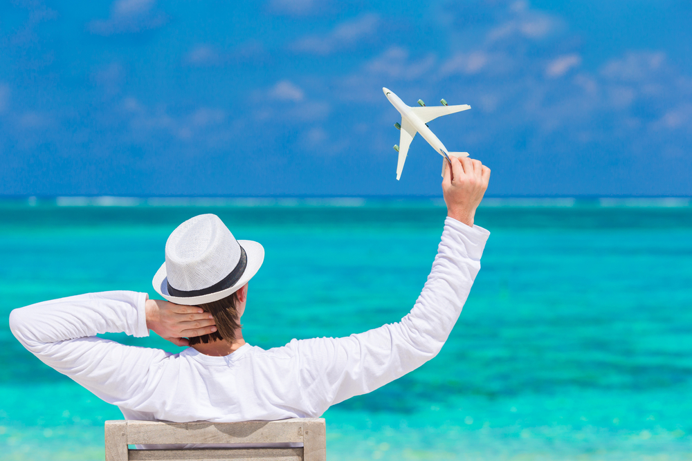 Ένας άντρας που φορά ένα λευκό καπέλο ενώ κάθεται σε μια ξύλινη καρέκλα σε μια παραλία, σηκώνοντας ένα αεροπλάνο παιχνίδι από πάνω. 