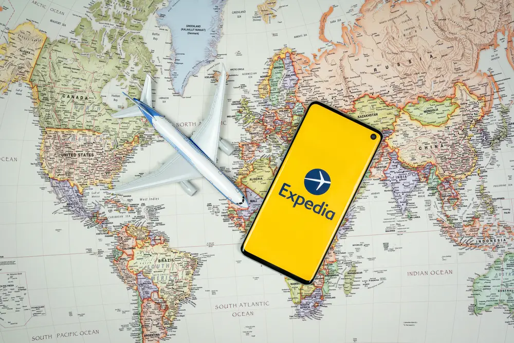 Smartphone που δείχνει το λογότυπο της εφαρμογής Expedia δίπλα σε ένα μοντέλο αεροπλάνου σε έναν παγκόσμιο χάρτη για να υποδείξει την έννοια της έμπνευσης ταξιδιού χρησιμοποιώντας ταξιδιωτικές εφαρμογές