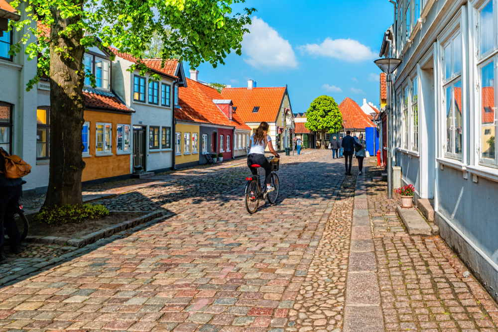 Πολύχρωμα σπίτια που απεικονίζονται την καλύτερη εποχή για να επισκεφθείτε τη Δανία, το καλοκαίρι, στο Odense με ανθρώπους να κάνουν ποδήλατο κατά μήκος του λιθόστρωτου δρόμου