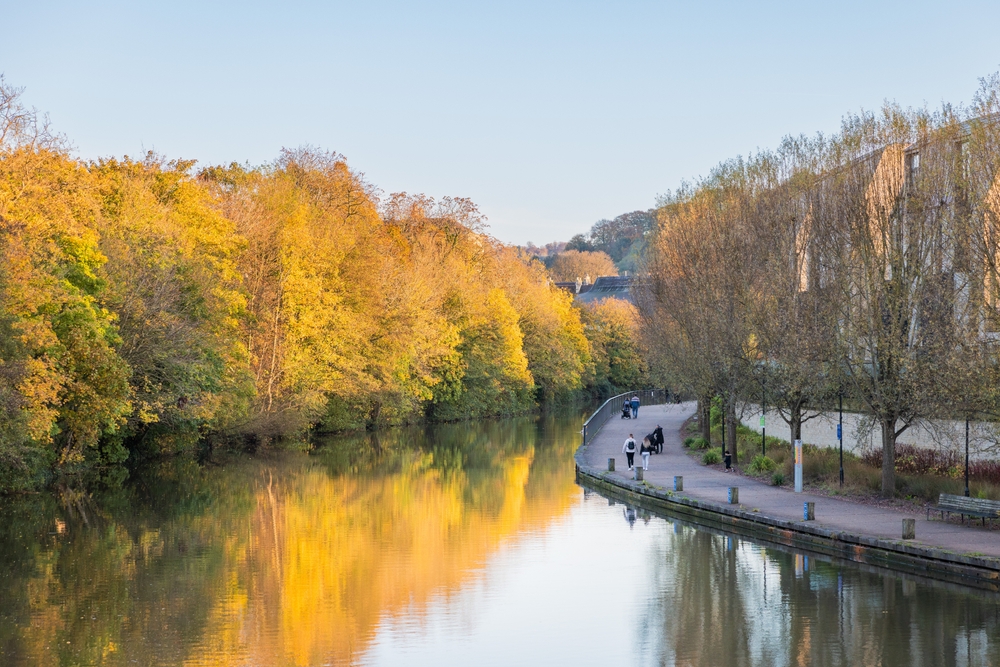 Θέα που αξίζει καρτ ποστάλ από χρυσά δέντρα και νερό με ανθρώπους που περπατούν κατά μήκος του ποταμού που απεικονίζεται κατά τη φθηνότερη περίοδο για να επισκεφθείτε το Ηνωμένο Βασίλειο, το φθινόπωρο