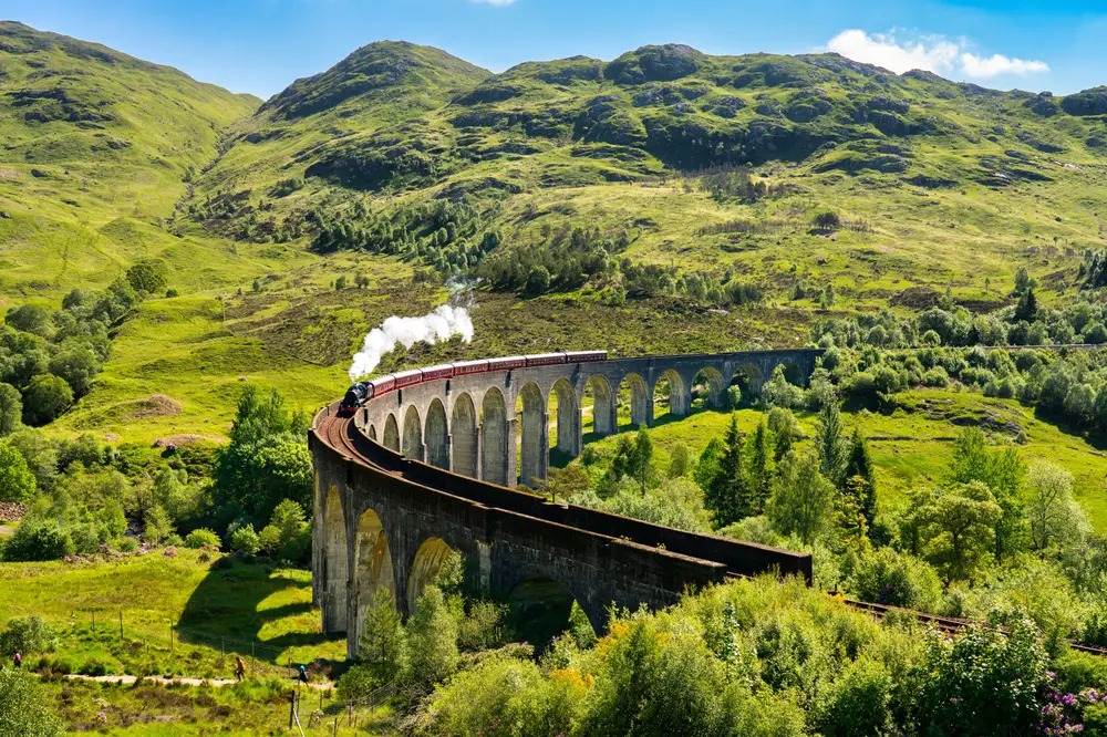 Φωτογραφημένο κατά τη διάρκεια της άνοιξης, τη συνολική καλύτερη εποχή για να επισκεφθείτε το Ηνωμένο Βασίλειο, το τρένο Glenfinnan Railway κάνει το δρόμο του πάνω από την οδογέφυρα και περιβάλλεται από πλούσια πράσινη βλάστηση