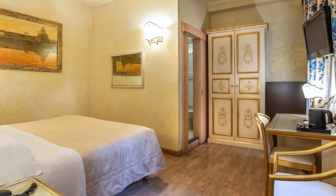 Ένα δωμάτιο ξενοδοχείου με ένα απλό διπλό κρεβάτι, ένα γραφείο, πίνακες ζωγραφικής της Φλωρεντίας στους απαλούς κίτρινους τοίχους και μια επιχρυσωμένη ντουλάπα στη γωνία στο Hotel Alba Palace στη Φλωρεντία, Ιταλία