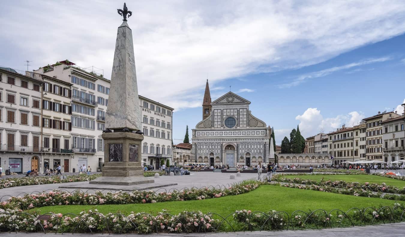 Μια πλατεία με λουλούδια και γρασίδι γύρω από ένα μνημείο και τη ζωγραφισμένη βασιλική Santa Maria Novella στο βάθος στη Φλωρεντία, Ιταλία