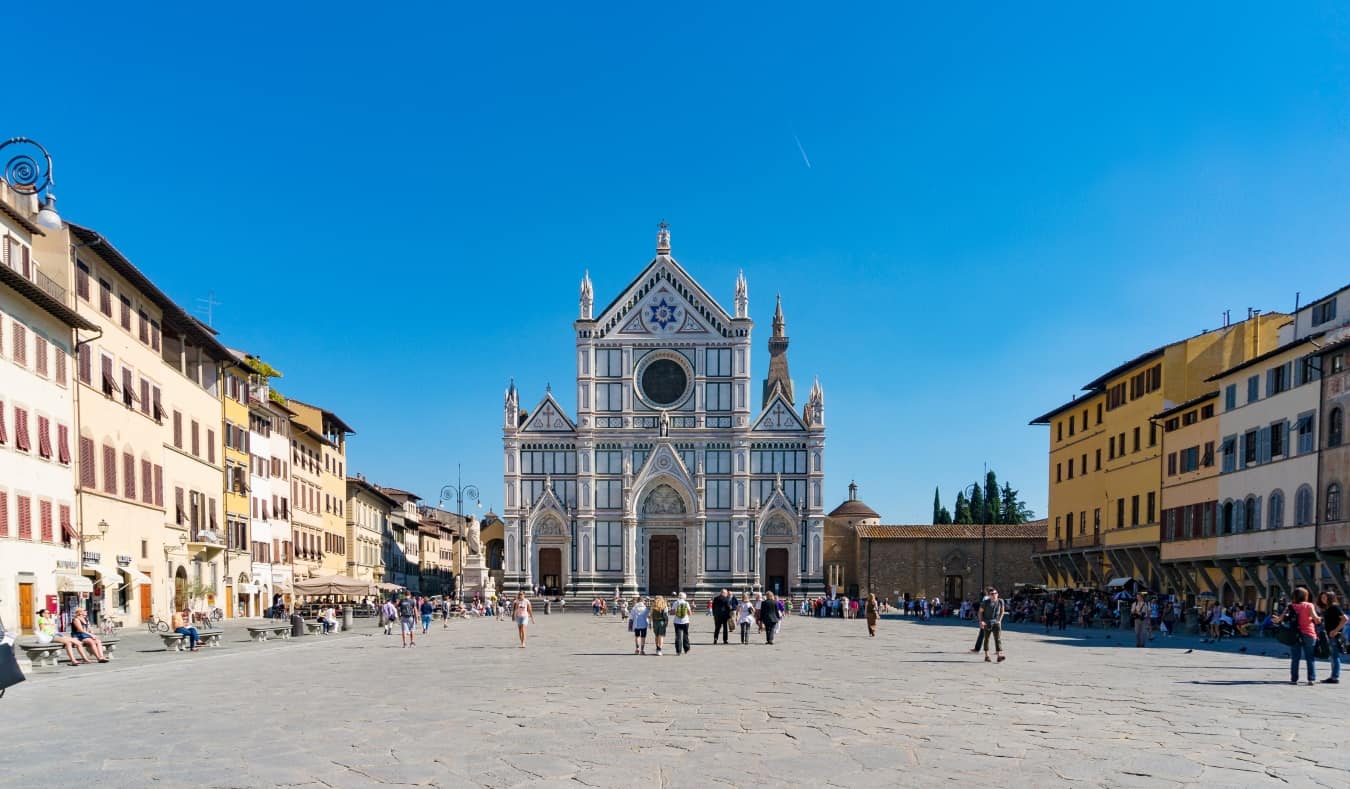 Μεγάλη πλατεία γεμάτη κτίρια, με ανθρώπους να περπατούν στον ανοιχτό χώρο μπροστά από τη μεγαλοπρεπή ζωγραφισμένη Βασιλική Santa Croce στη Φλωρεντία της Ιταλίας