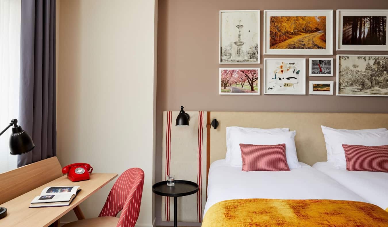 Ένα σύμπλεγμα από φωτογραφίες και σχέδια κρέμεται πάνω από ένα κρεβάτι με κόκκινα μαξιλάρια και πορτοκαλί κουβέρτες, δίπλα σε ένα ξύλινο γραφείο με ένα κόκκινο περιστροφικό τηλέφωνο πάνω του στο Hotel Indigo Madrid-Princesa στη Μαδρίτη, Ισπανία