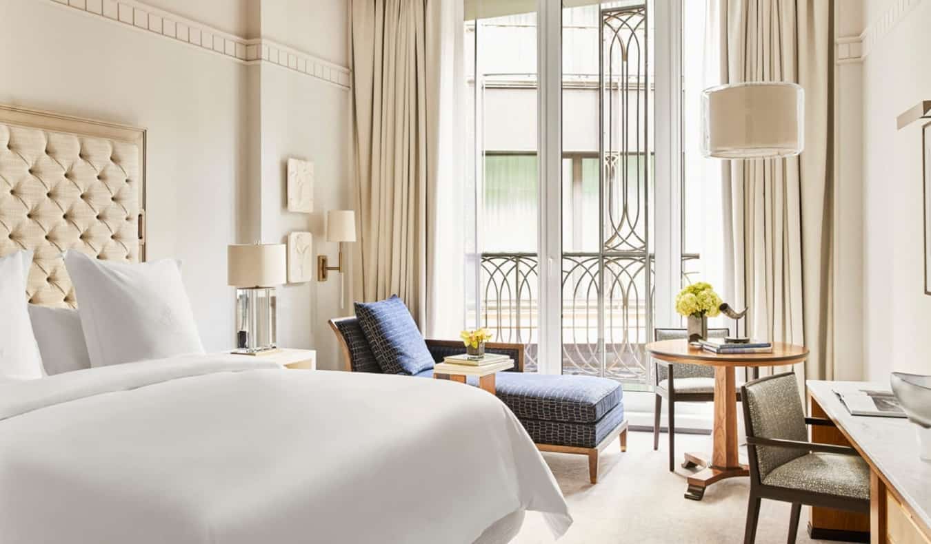 Ένα πολυτελές δωμάτιο ξενοδοχείου σε κρεμ χρώμα με ένα μεγάλο κρεβάτι, μπλε σεζλόν δίπλα στο ανοιχτό παράθυρο και ένα γραφείο στο Four Seasons στη Μαδρίτη, Ισπανία